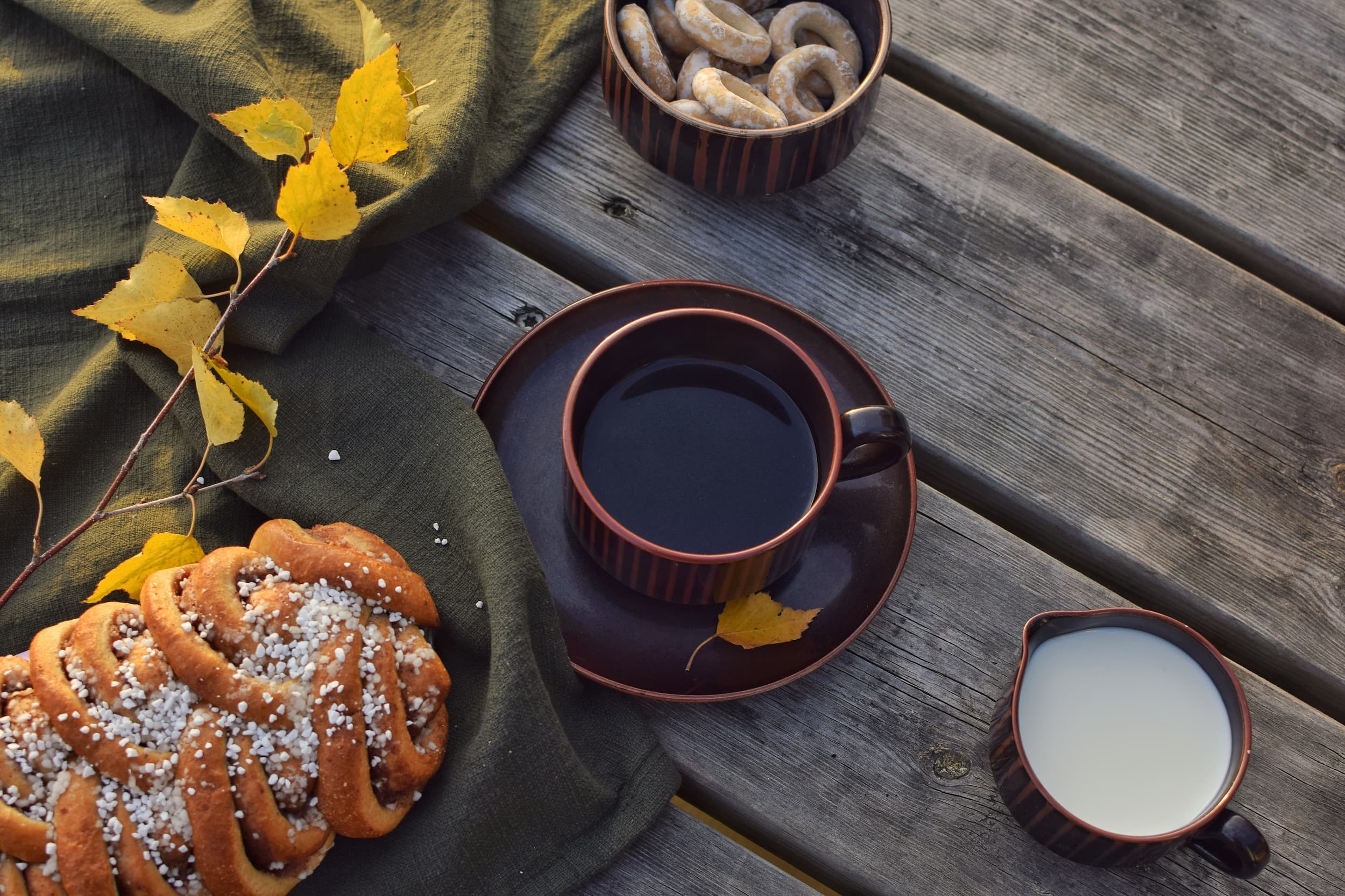 Kaffee mit Gebäck im herbstlichen Ambiente Tasse Kaffee mit Milch und Zimtschnecke auf Holztisch, dekoriert mit Herbstlaub.