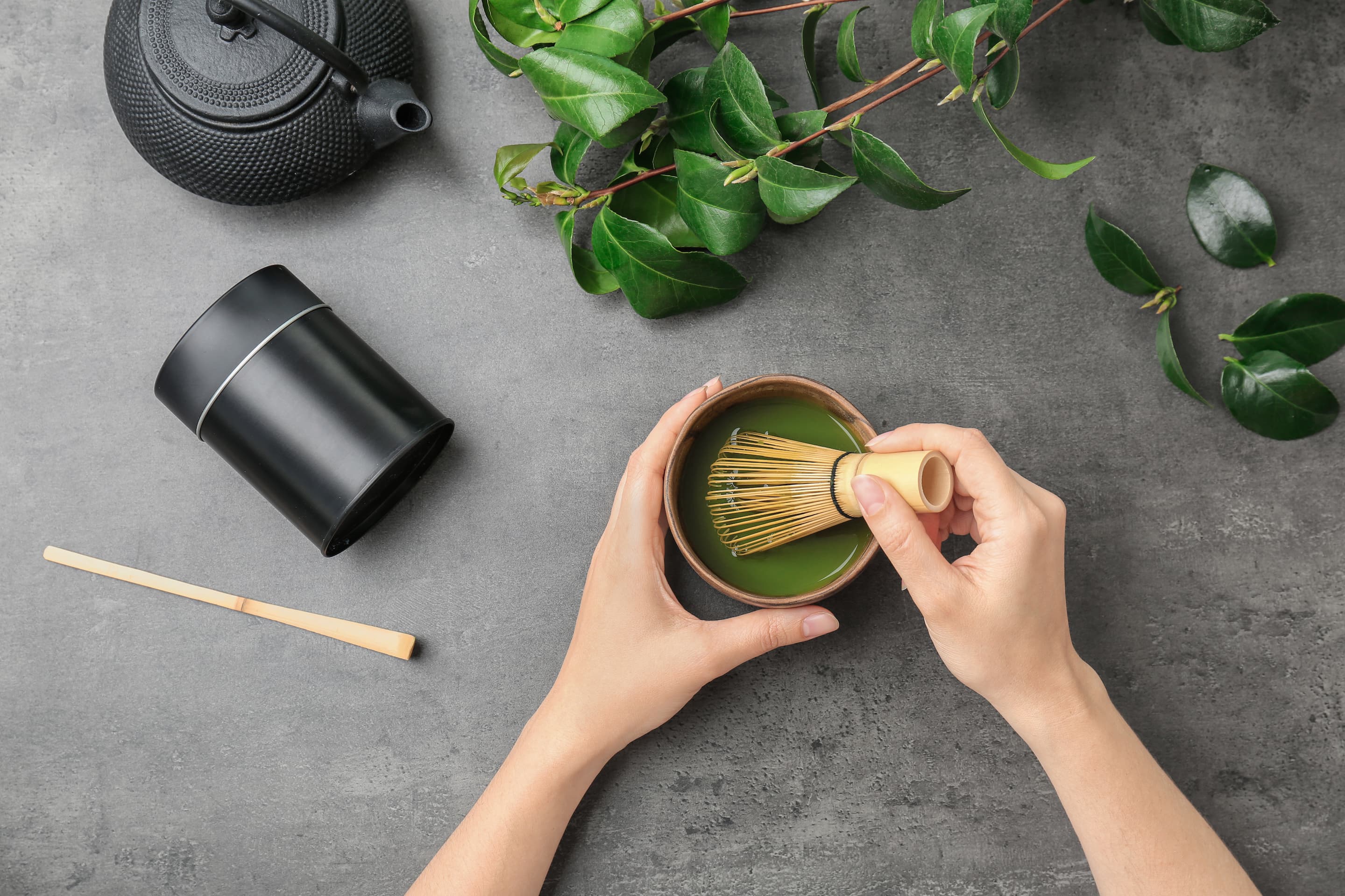 Traditionelle Matcha Tee Zubereitung Hände bereiten Matcha Tee mit Bambusbesen in Schale zu, Teekanne und Pflanzen daneben.