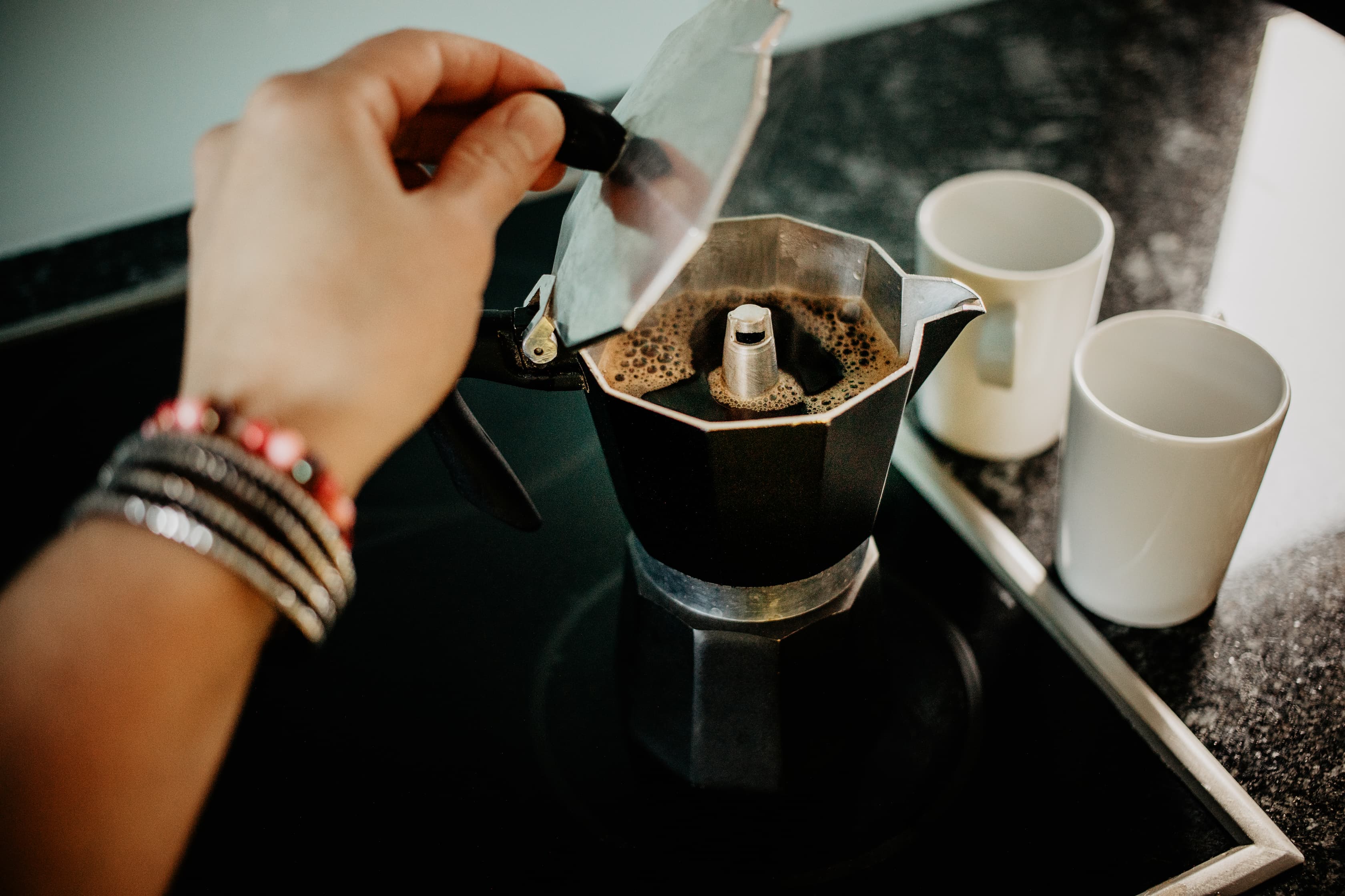 Espressozubereitung im Espressokocher Hand öffnet Espressokocher auf Herd, frischer Kaffee steigt auf, zwei weiße Tassen daneben.