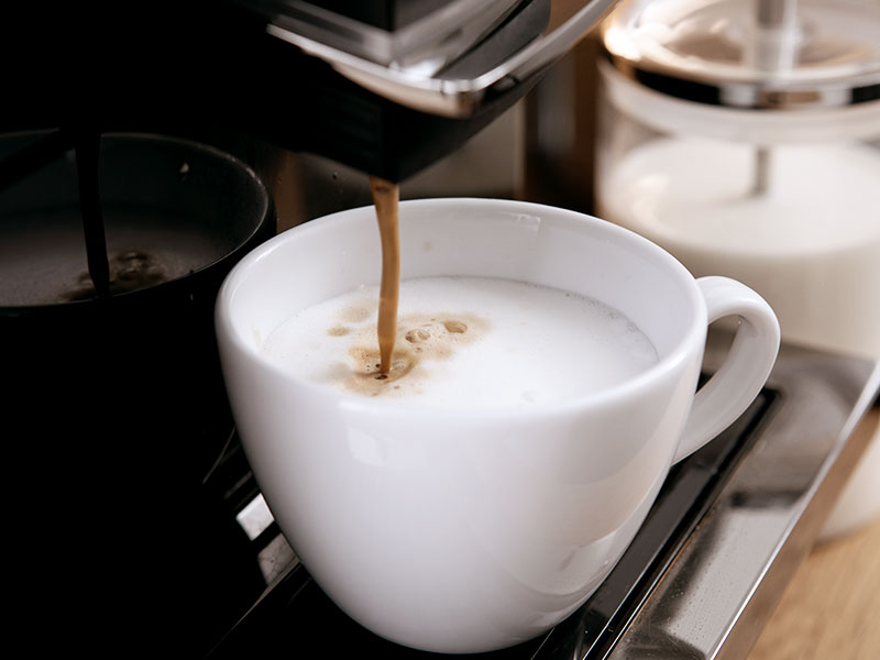 MP10 Kaffeevollautomat bereitet Cappuccino mit Milchschaum in einer weißen Tasse zu, Milchbehälter im Hintergrund sichtbar.