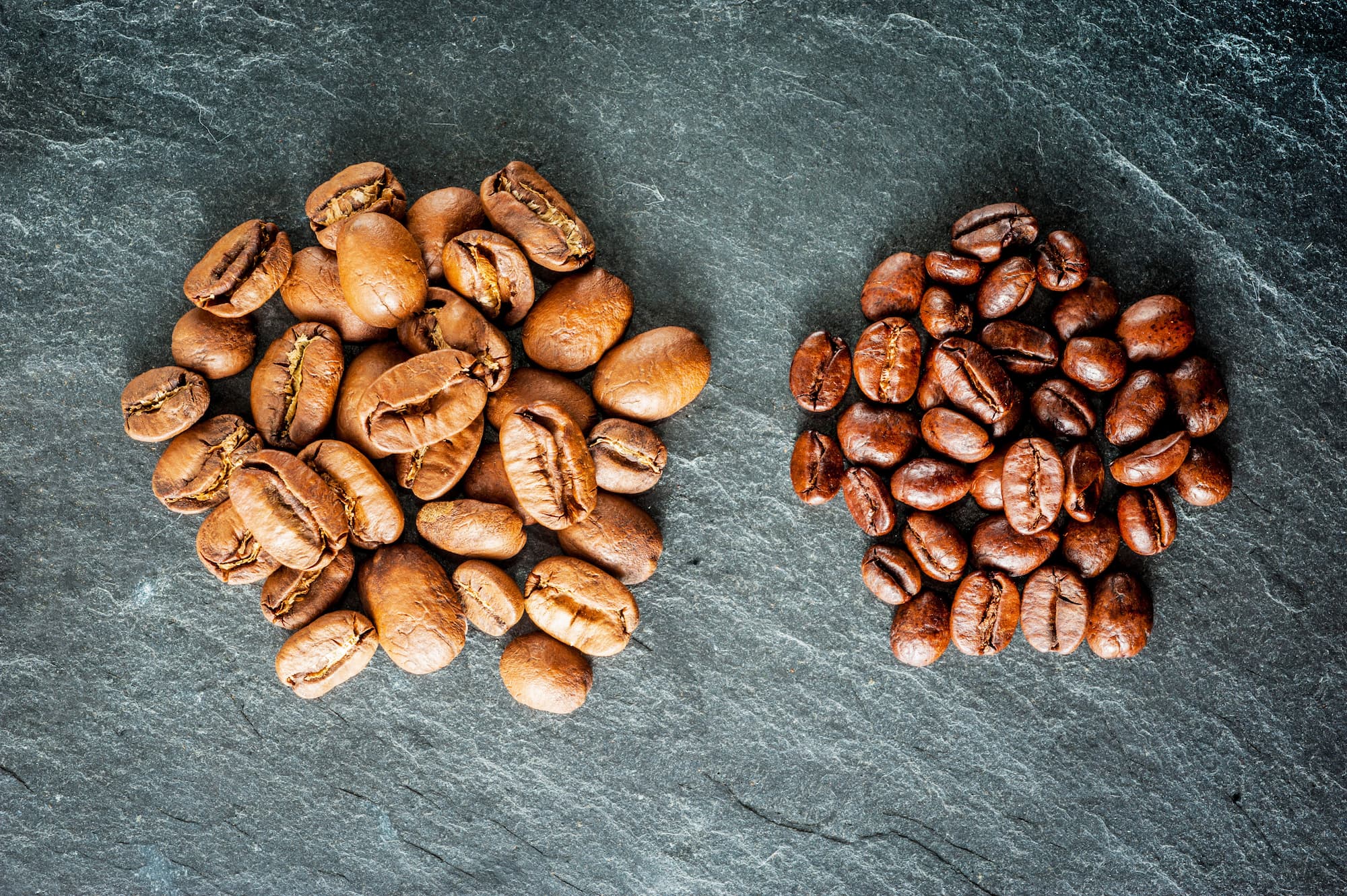 Unterschied zwischen heller und dunkler Kaffeeröstung Zwei Häufchen Kaffeebohnen – links helle Röstung, rechts dunkle Röstung auf Steinplatte.