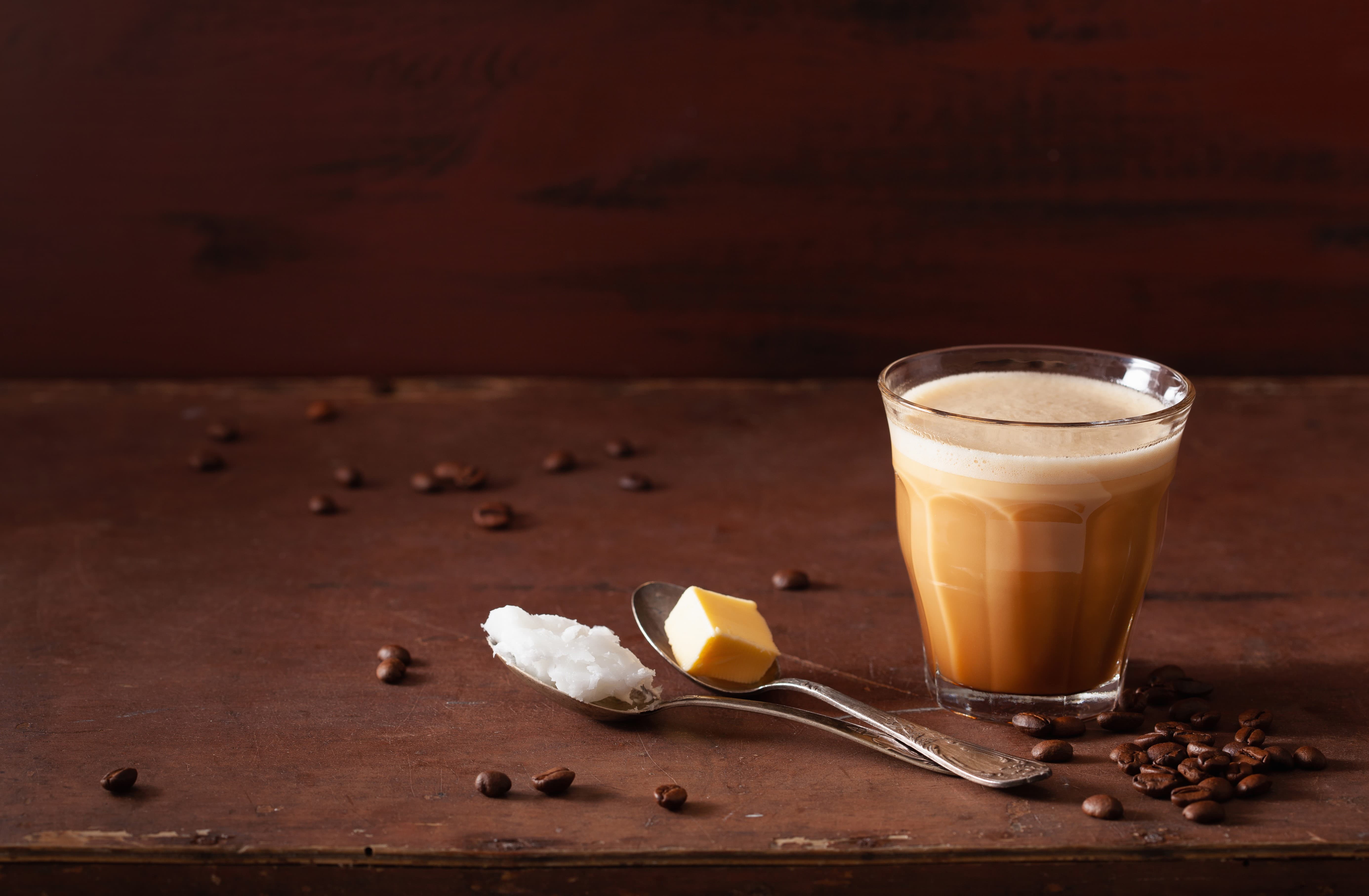 Glas mit Bulletproof Coffee, daneben Butter und Kokosoel auf einem Loeffel, dekoriert mit Kaffeebohnen.