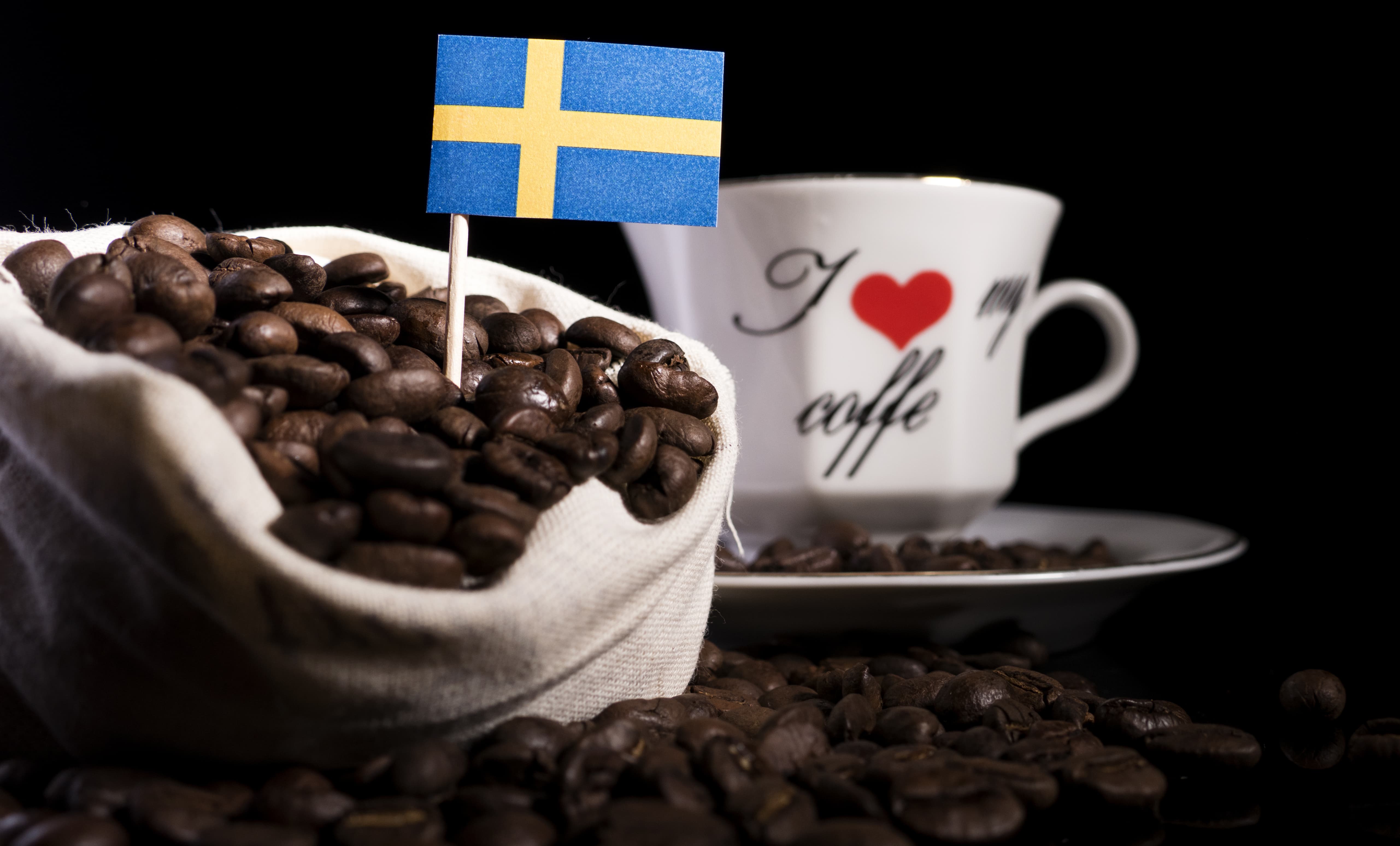 Kaffeebohnen mit schwedischer Flagge Sack voller Kaffeebohnen mit schwedischer Flagge, im Hintergrund eine weiße Tasse mit Aufschrift.