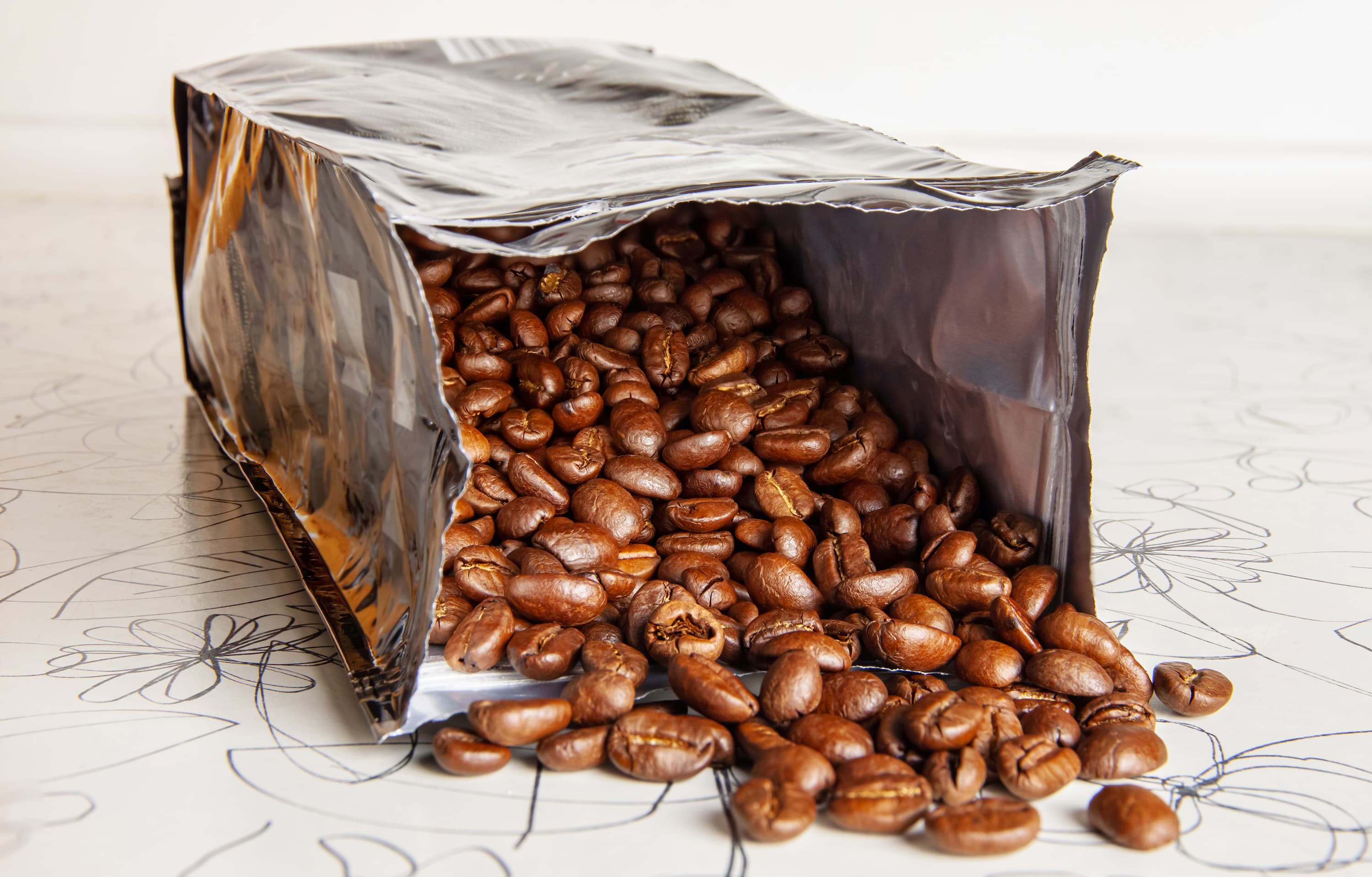 Geöffnete Kaffeeverpackung mit Bohnen Silberne Kaffeeverpackung, aus der frische Kaffeebohnen herausfallen.