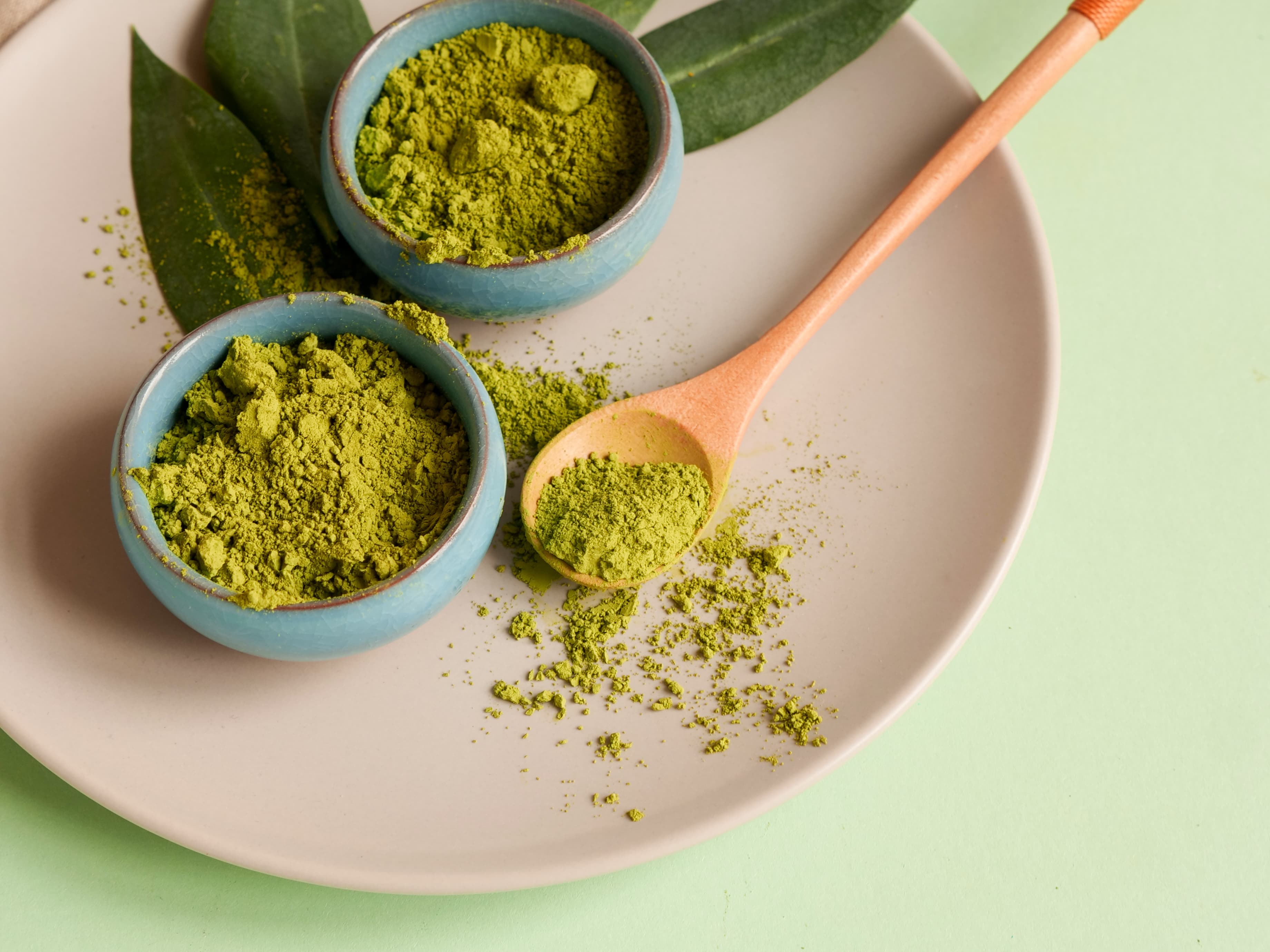 Zwei Schalen mit gruenem Matcha-Pulver und Holzloeffel auf hellgruenem Untergrund.