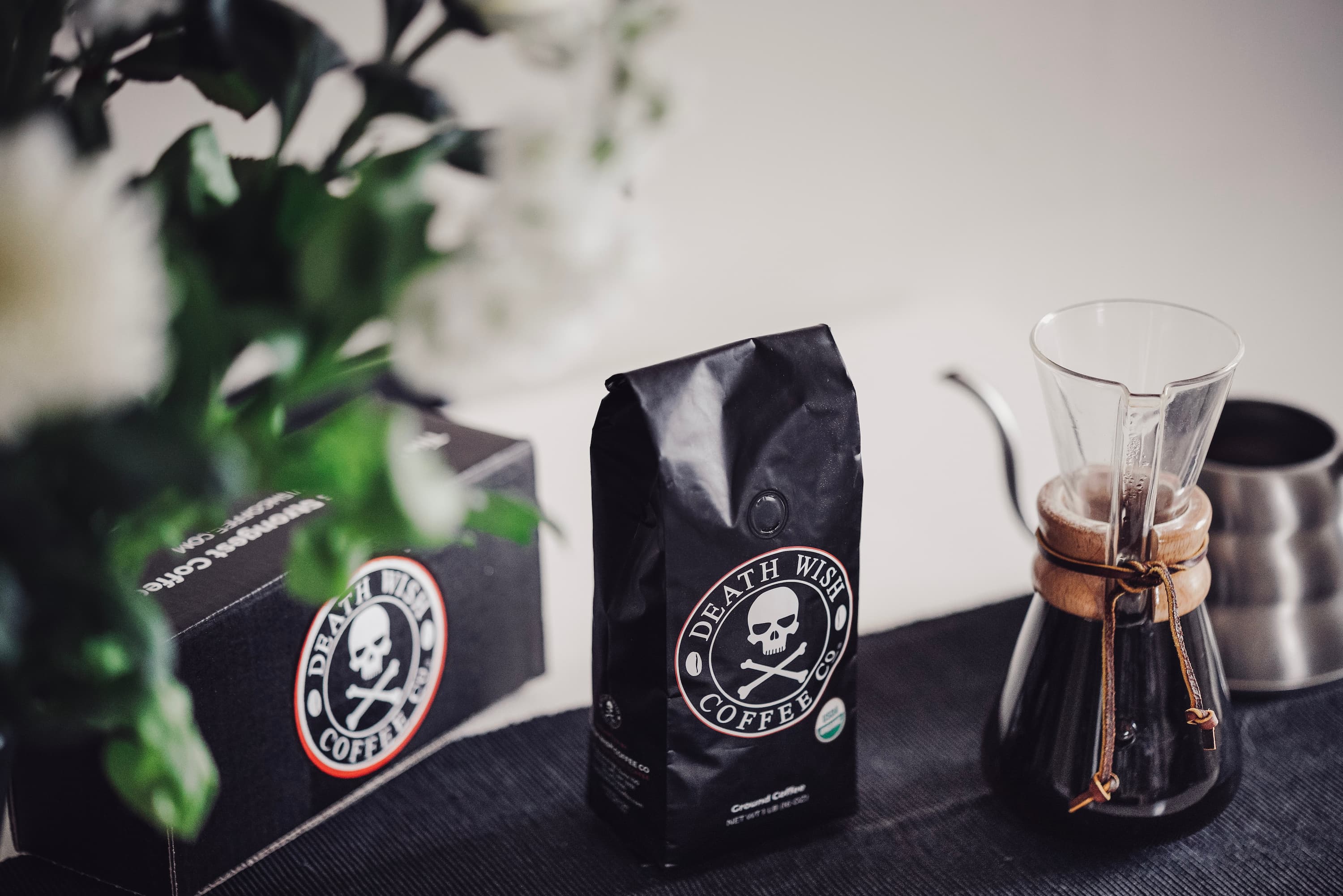 Schwarze Death Wish Coffee Packung neben Chemex Kaffeekanne und Karton, Blumen im Vordergrund.