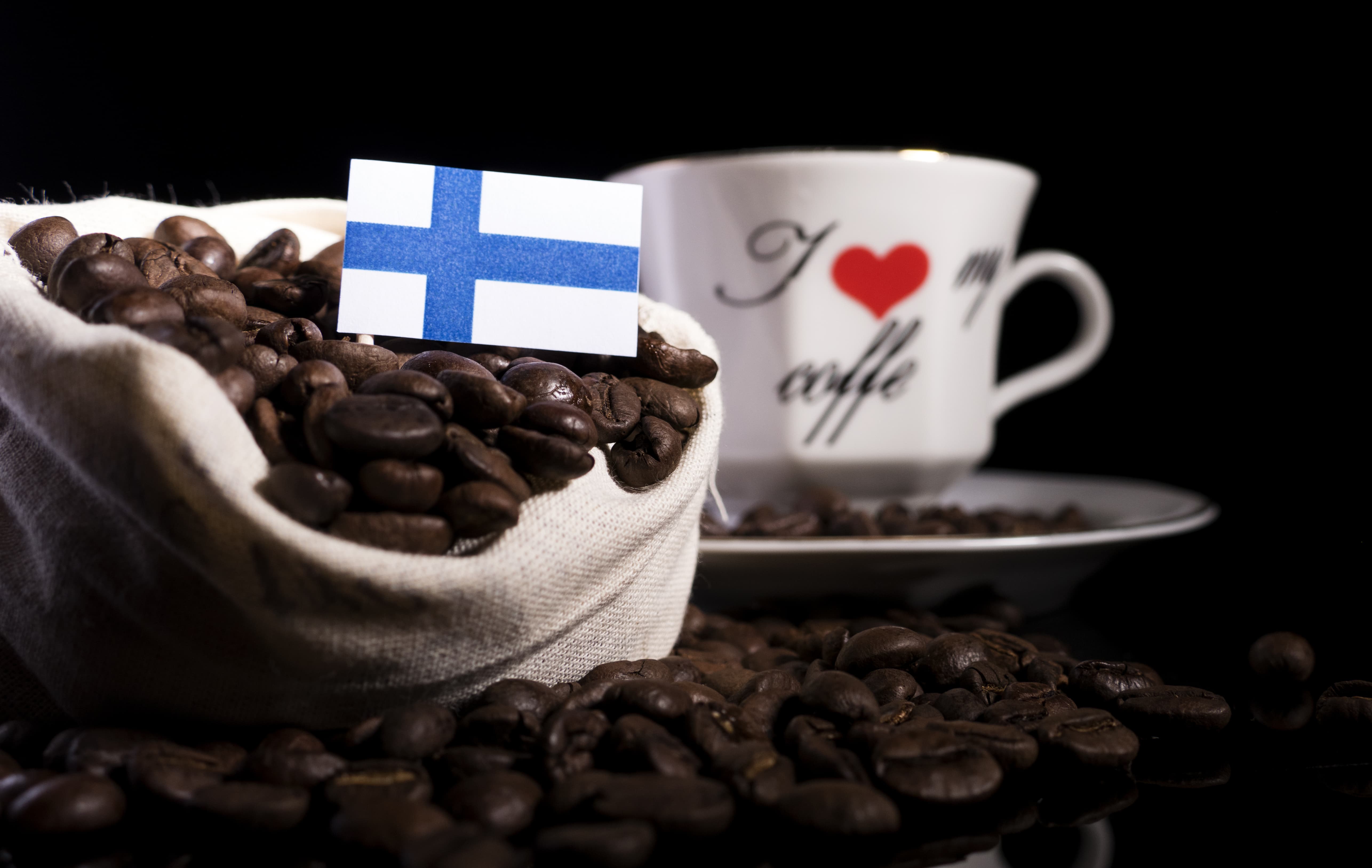 Kaffeebohnen mit finnischer Flagge Sack mit Kaffeebohnen, darin kleine finnische Flagge, im Hintergrund eine Kaffeetasse.