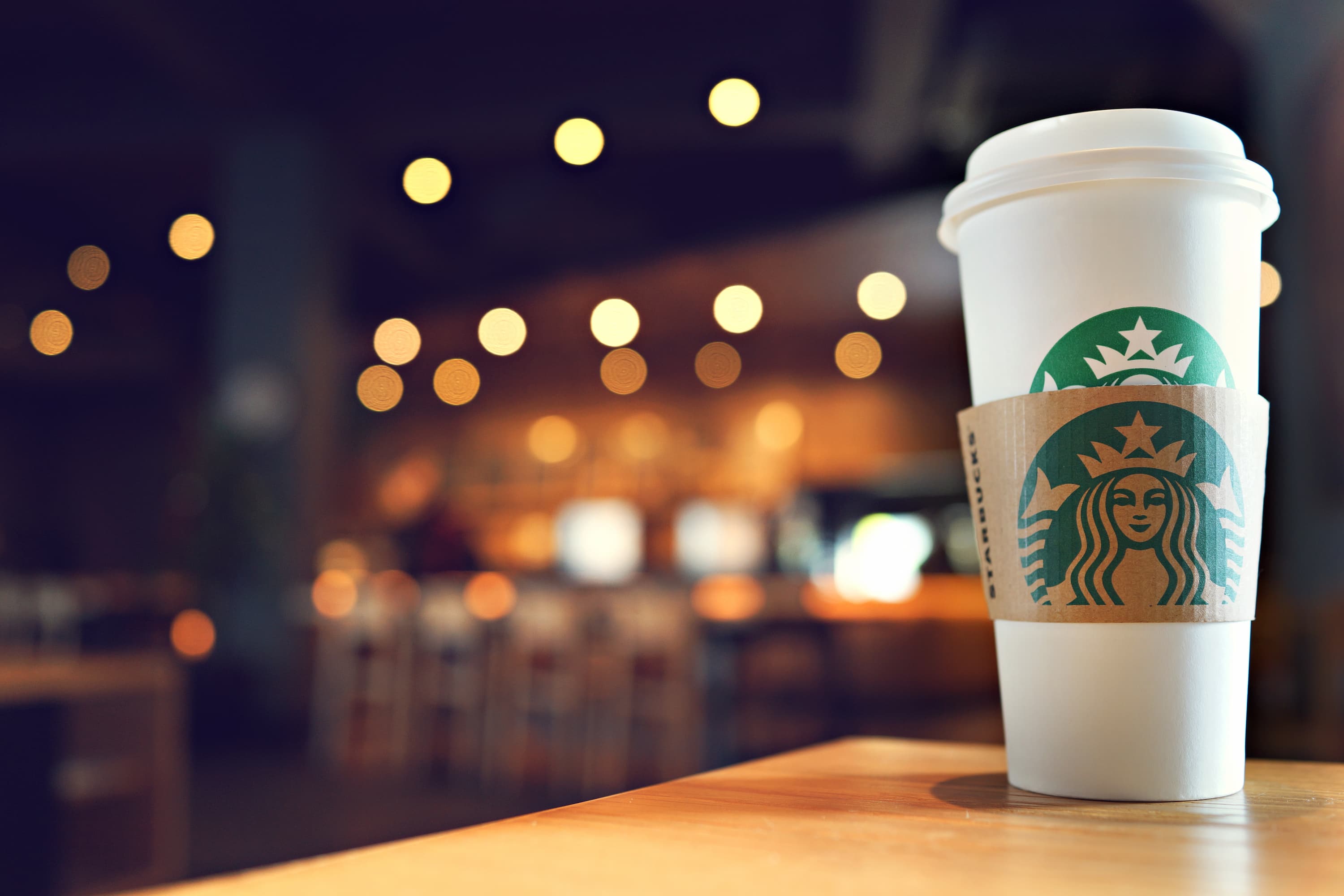 Weißer Starbucks Pappbecher mit Logo auf Holztisch, unscharfer Café-Hintergrund mit Lichtern.