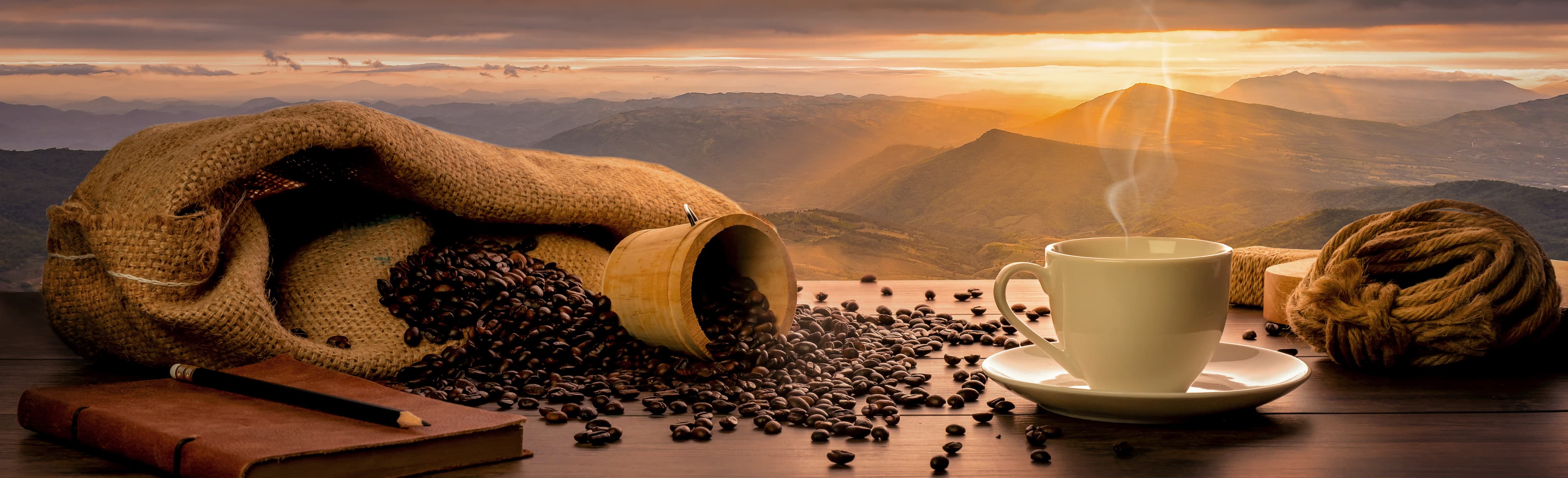 Kaffeetasse und Bohnen bei Sonnenaufgang Weiße Kaffeetasse mit Dampf neben Kaffeesack und Bohnen vor Sonnenaufgang über Berglandschaft.