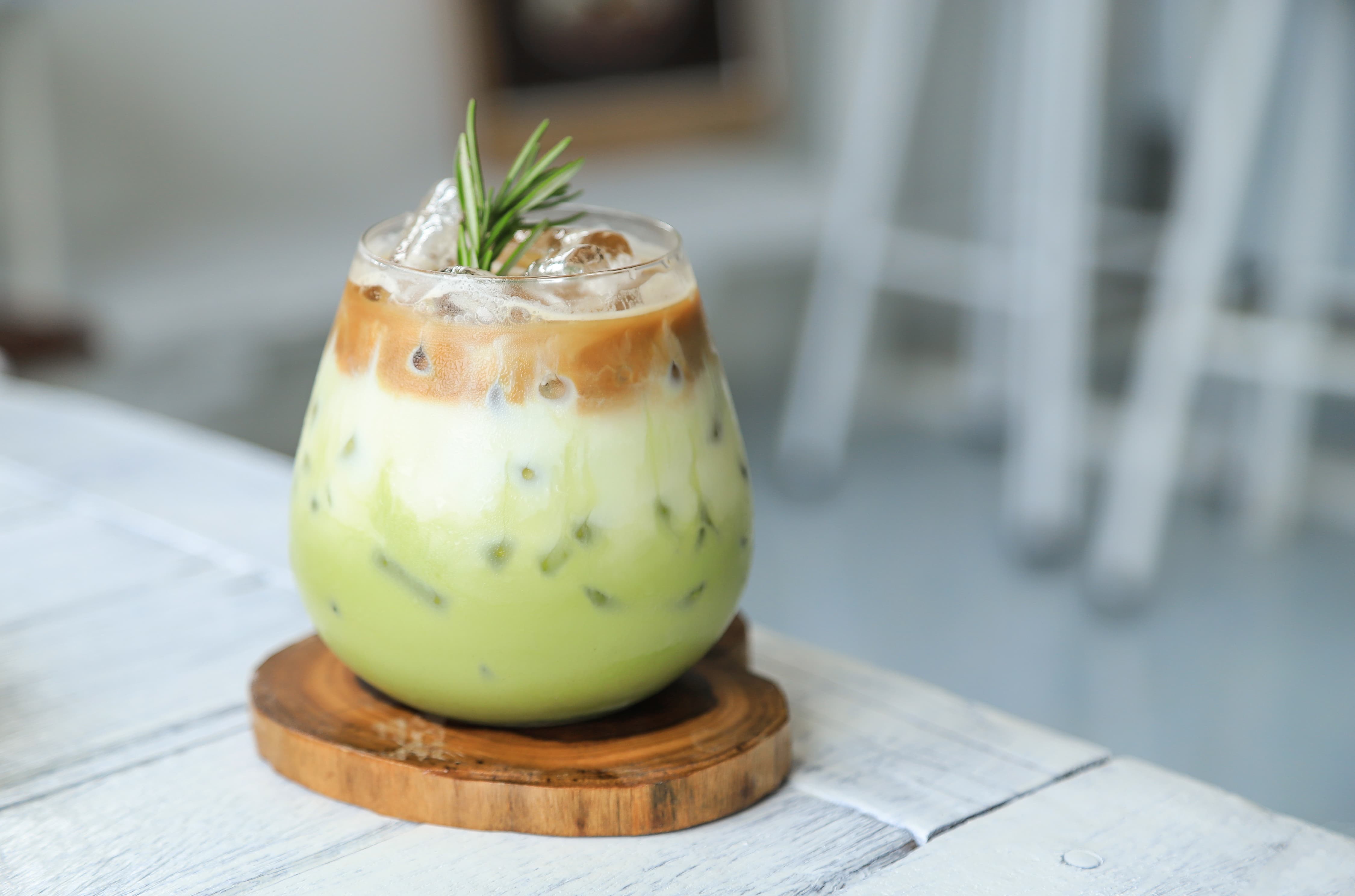 Iced Matcha Latte mit Kaffee Glas mit Iced Matcha Latte, Schichten aus grünem Tee, Milch und Kaffee, dekoriert mit Rosmarin.